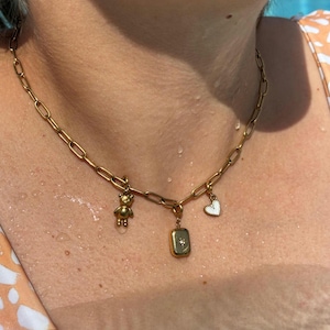 Könnte beinhalten: Goldfarbene Kette mit drei Anhängern. Die Anhänger umfassen einen kleinen Teddybären, einen rechteckigen Anhänger mit einem kleinen Edelstein und ein weißes Herz. Die Halskette wird am Hals einer Person getragen.