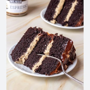 May include: A slice of chocolate cake with a light-colored filling and chocolate frosting on a white plate. A fork is inserted into the cake. Another slice of cake is visible in the background. A jar of espresso is partially visible.
