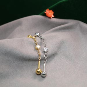 May include: Two belly button rings are shown, one gold-toned and one silver-toned. Each has a chain with a clear, round gem and a dangling ball. The jewellery is set against a grey fabric background.