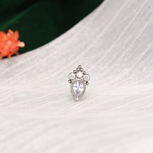Pode incluir: Uma joia corporal de metal prateado com uma pedra preciosa transparente em forma de pera no centro, encimada por três pedras redondas mais pequenas dispostas em leque, acentuadas por pequenas esferas metálicas entre as pedras.