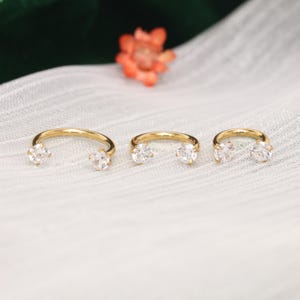 Puede incluir: Tres anillos dorados en forma de herradura, cada uno con una piedra preciosa transparente de corte redondo en ambos extremos. Las bandas de metal son lisas y pulidas, con las piedras sujetas por engastes de cuatro puntas.