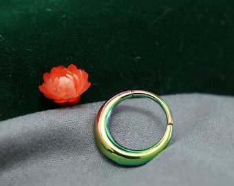 May include: A hinged segment ring with a rainbow finish. The jewellery piece displays a spectrum of colours, including green, blue, and gold. The ring is placed on a grey surface with a small orange flower in the background.