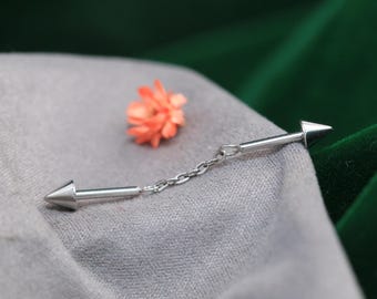 May include: A silver industrial barbell earring with cone-shaped ends, connected by a delicate chain. The jewelry is displayed on a gray fabric with a small orange flower in the background. The earring is a stylish accessory.