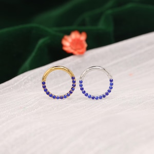 May include: Two circular hoop rings, one in a gold tone and one in a silver tone, each featuring a row of small, vibrant blue circular gemstones embedded along the lower half of the hoop, designed with a hinged segment for closure.