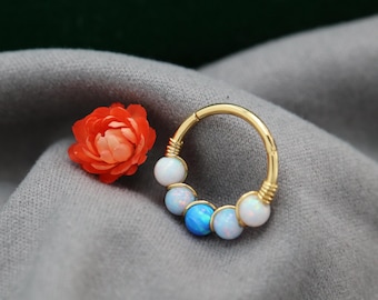 May include: A gold septum ring featuring a row of white and blue opal beads. The ring is circular with a hinged design. An orange flower is placed beside the ring on a grey surface.