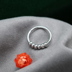 May include: A silver nose ring with a hinged closure. The ring has six small, round beads along the bottom curve. An orange flower is in the lower left corner, against a grey fabric background.