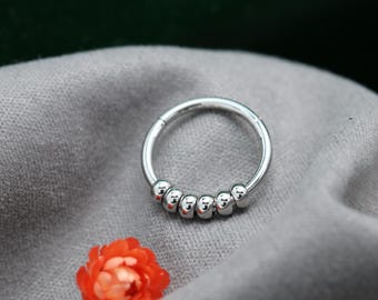May include: A silver nose ring with a hinged closure. The ring has six small, round beads along the bottom curve. An orange flower is in the lower left corner, against a grey fabric background.