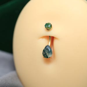 Puede incluir: Un piercing para el ombligo con una barra curva dorada. La gema superior es una piedra verde pequeña y redonda. La gema inferior es una piedra verde más grande en forma de lágrima con inclusiones más oscuras.