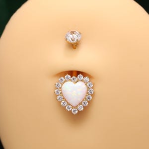 Op de afbeelding: Een gouden navelpiercing met een hartvormige witte opaalsteen omringd door een halo van kleine heldere, rondgeslepen kristallen. Het bovenste deel is een enkel helder rondgeslepen kristal in goud op een glad, huidkleurig oppervlak.