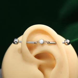 May include: A silver industrial barbell earring with a white opal centre stone and two silver ball ends. The earring is shown on a mannequin ear. The jewellery is designed for ear piercings.