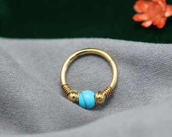May include: A gold-coloured nose ring featuring a turquoise bead. The ring has a circular design with coiled wire detailing on either side of the bead. The background is a soft grey fabric.