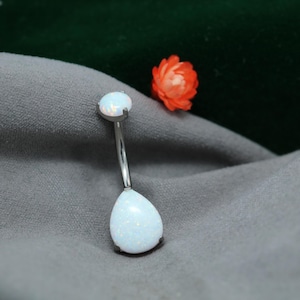 May include: A silver belly button ring with two white opal stones. The top stone is round, and the bottom stone is teardrop-shaped. The ring is on a gray surface with a small orange flower in the background.