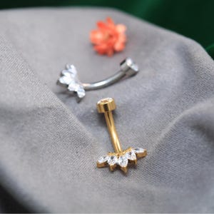 Peut inclure: Deux piercings de nombril. L'un est doré avec un groupe de cristaux transparents et un seul cristal supérieur. L'autre est argenté avec un groupe de cristaux. Une petite fleur orange est en arrière-plan. Les bijoux sont sur une surface grise.