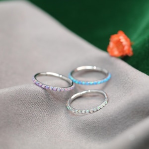 Puede incluir: Tres anillos de plata con una banda de pequeños ópalos de colores. Un anillo tiene una banda de ópalos morados, otro de ópalos azules y el tercero de ópalos blancos. Los anillos se exhiben sobre una tela gris con un fondo verde y naranja.