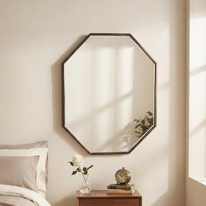 May include: An octagonal wall mirror with a dark metal frame hanging above a wooden bedside table. The table features a single drawer with a small knob, four tapered legs, and holds a glass vase with a white rose and a vintage-style clock.