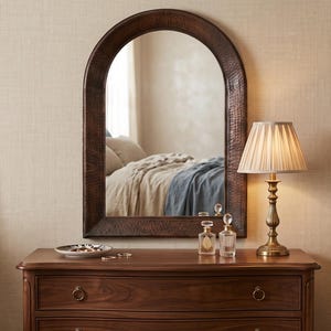 May include: An arched wall mirror with a dark brown, hammered metal frame. The mirror is mounted above a wooden dresser, reflecting a bedroom interior. The frame features a textured, dimpled surface that catches the light, providing a distinct contrast to the smooth, polished wood of the dresser below it.