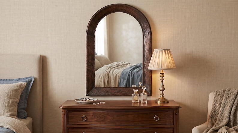 May include: An arched wall mirror with a dark brown, hammered metal frame. The mirror is mounted above a wooden dresser, reflecting a bedroom interior. The frame features a textured, dimpled surface that catches the light, providing a distinct contrast to the smooth, polished wood of the dresser below it.