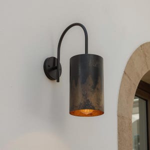 Puede incluir: Aplique de pared exterior negro con una pantalla cilíndrica y un brazo curvo. La pantalla tiene un acabado oscuro texturizado con detalles dorados. El aplique está montado en una pared blanca.