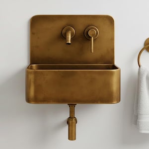 May include: A wall-mounted, antique brass sink with a rectangular basin and a matching faucet and handle. A brass towel ring holds a white towel. The sink is mounted on a white wall.