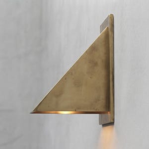 May include: A triangular wall-mounted light fixture crafted from brushed brass. The fixture features a flat rectangular backplate attached to the wall, with a slanted triangular shade extending outward, casting a warm glow downward from the base.