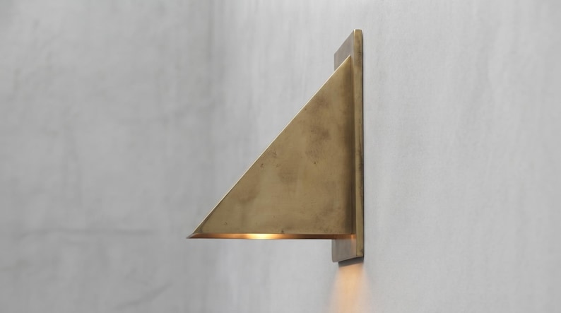 May include: A triangular wall-mounted light fixture crafted from brushed brass. The fixture features a flat rectangular backplate attached to the wall, with a slanted triangular shade extending outward, casting a warm glow downward from the base.