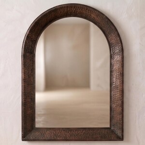 May include: An arched wall mirror featuring a dark, hammered copper frame with a textured, metallic finish. The mirror has a rounded top and straight sides, providing a clear reflective surface that captures the surrounding light and space.