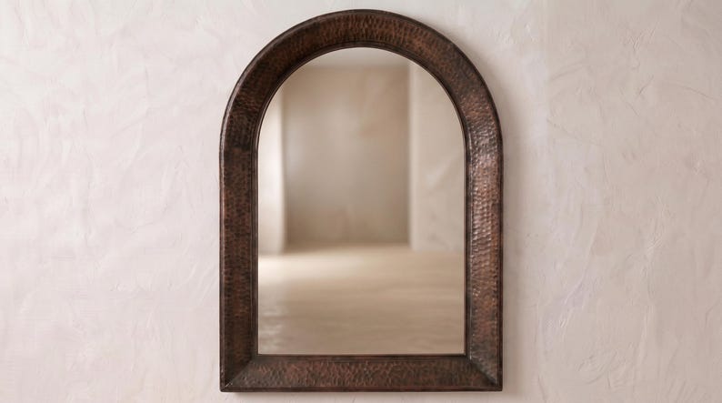 May include: An arched wall mirror featuring a dark, hammered copper frame with a textured, metallic finish. The mirror has a rounded top and straight sides, providing a clear reflective surface that captures the surrounding light and space.