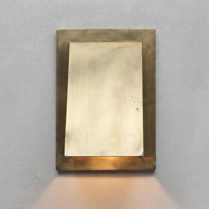 May include: A rectangular wall sconce made of brushed brass with a flat, vertical plate centered over a slightly larger rectangular base. The fixture emits a warm, downward-facing light that illuminates the area directly beneath the metal structure.