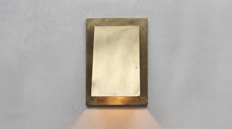 May include: A rectangular wall sconce made of brushed brass with a flat, vertical plate centered over a slightly larger rectangular base. The fixture emits a warm, downward-facing light that illuminates the area directly beneath the metal structure.