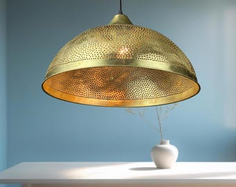 Puede incluir: Una lámpara colgante dorada en forma de cúpula con un diseño perforado. La luz cuelga de un cable negro e ilumina una mesa blanca. Un pequeño jarrón blanco con una ramita de flores secas está sobre la mesa.