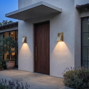 May include: A modern home exterior featuring a dark wood front door with a vertical handle, flanked by two brass-toned wall sconces that cast a warm downward glow. The building has a light-colored stucco facade and large black-framed windows.