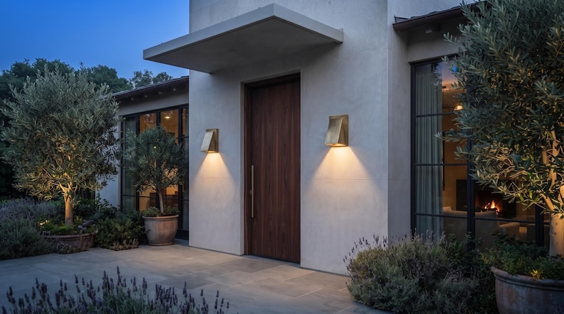 May include: A modern home exterior featuring a dark wood front door with a vertical handle, flanked by two brass-toned wall sconces that cast a warm downward glow. The building has a light-colored stucco facade and large black-framed windows.