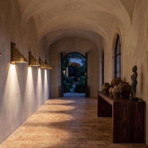 May include: A long hallway with a vaulted ceiling and stone floor features a series of triangular brass wall sconces that cast downward light. A dark wood console table holds several round ceramic vessels and a small dark statue near a window.