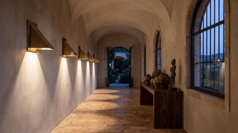 May include: A long hallway with a vaulted ceiling and stone floor features a series of triangular brass wall sconces that cast downward light. A dark wood console table holds several round ceramic vessels and a small dark statue near a window.
