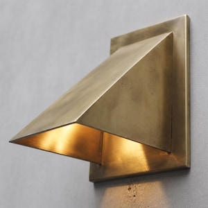 Puede incluir: Un aplique de pared con una placa trasera cuadrada y una pantalla triangular inclinada hacia abajo hecha de latón cepillado. La lámpara emite un brillo cálido y dorado desde debajo de la pantalla, iluminando la superficie bajo la unidad.