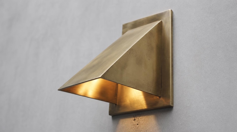 May include: A wall-mounted light fixture with a square backplate and a triangular, downward-angled shade made of brushed brass. The fixture emits a warm, golden glow from beneath the shade, illuminating the surface directly below the unit.