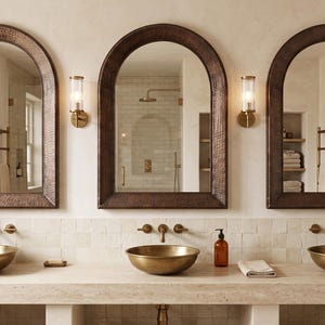 May include: Three arched wall mirrors with dark, hammered metal frames are mounted above a long stone vanity. Each mirror is flanked by a cylindrical wall sconce. Three brass vessel sinks sit on the vanity, paired with wall-mounted faucets.