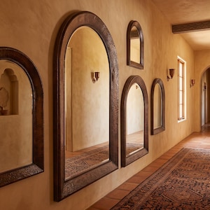 May include: A series of arched mirrors with dark, textured metal frames mounted on a warm-toned wall. The mirrors vary in size and shape, reflecting the hallway space. The arrangement includes several wall-mounted light fixtures with metallic finishes.