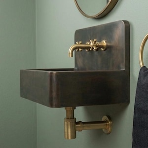 Puede incluir: Un lavabo rectangular de bronce oscuro montado en la pared con un panel trasero alto, que cuenta con un grifo de latón con dos mandos en cruz. Debajo del lavabo hay un sifón de latón. Un toallero de aro de latón sostiene una toalla gris oscuro.
