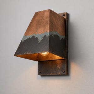 May include: A wall-mounted light fixture featuring a trapezoidal shade with a dual-tone finish. The upper portion is a warm copper color, while the lower section is a dark, weathered charcoal gray with a textured, uneven transition line.