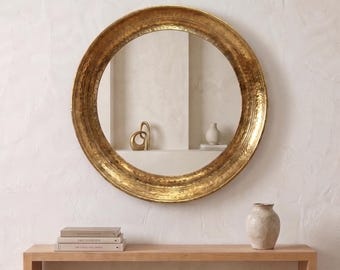 May include: A round wall mirror featuring a wide, textured gold frame with a hammered metal finish. The mirror reflects a room interior with a small decorative sculpture and a vase. The mirror is mounted on a plain, light-colored wall.
