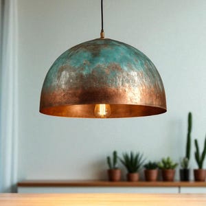 May include: A dome-shaped pendant light with a copper and teal finish. The interior of the lamp is copper, and it hangs from a black cord. A single lightbulb is visible inside the lamp. The light fixture is suspended above a wooden surface.