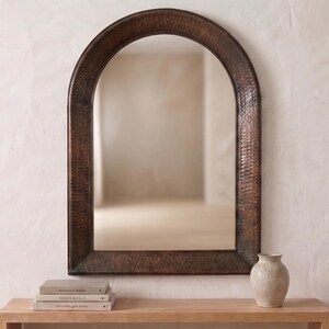 May include: An arched wall mirror featuring a dark, textured, hammered metal frame with a deep bronze finish. The mirror has a rounded top and straight sides, reflecting the surrounding space within its clear, reflective glass surface.