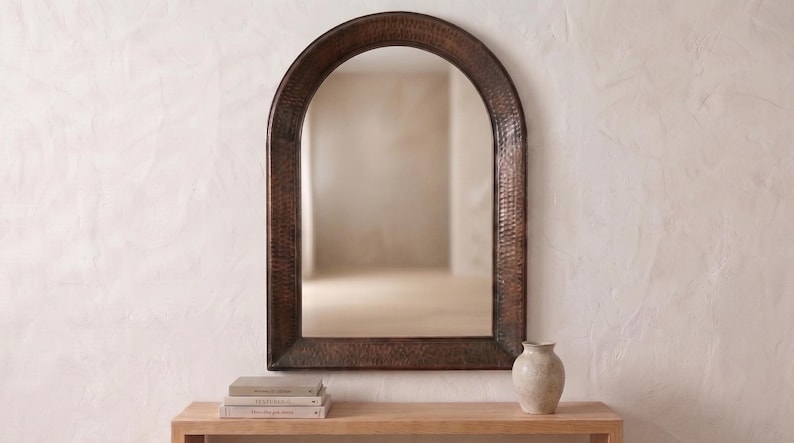 May include: An arched wall mirror featuring a dark, textured, hammered metal frame with a deep bronze finish. The mirror has a rounded top and straight sides, reflecting the surrounding space within its clear, reflective glass surface.
