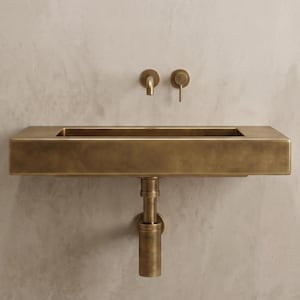 Könnte beinhalten: Ein rechteckiges Wandwaschbecken mit gebürstetem Messing-Finish. Das Waschbecken verfügt über ein passendes Messing-Abflussrohr, das nach unten verläuft, sowie eine wandmontierte Messingarmatur mit einem Hebelgriff direkt über dem Becken.