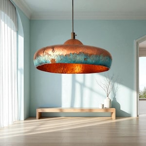 May include: A large, copper-coloured pendant light fitting with a distressed teal band around the edge. The interior of the light is a warm orange-gold colour. The fitting hangs from a dark cord in a room with light blue walls.