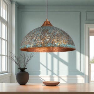 May include: A dome-shaped pendant light with a copper finish and a distressed blue-green patina. The light fixture hangs from a black cord and is suspended above a wooden table. The light has a perforated design and is a decorative element.
