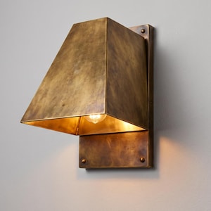May include: A wall-mounted sconce light fixture crafted from aged brass with a trapezoidal shade. The fixture features a rectangular backplate secured with visible rivets and an open bottom design that emits a warm glow from an internal bulb.