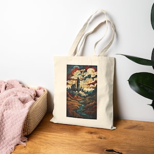 May include: A cream tote bag displaying a landscape painting. The artwork shows a castle with tall spires, a winding river, and colourful clouds in shades of red, orange, and blue. The bag has long handles.