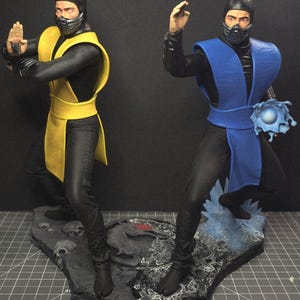 Könnte beinhalten: Zwei Actionfiguren auf einem Sockel mit einem Rasterlineal. Die linke Figur trägt einen gelben Überwurf über einem schwarzen Anzug mit Gesichtsmaske. Die rechte Figur trägt einen blauen Überwurf über einem schwarzen Anzug und hält eine blaue Energiekugel.